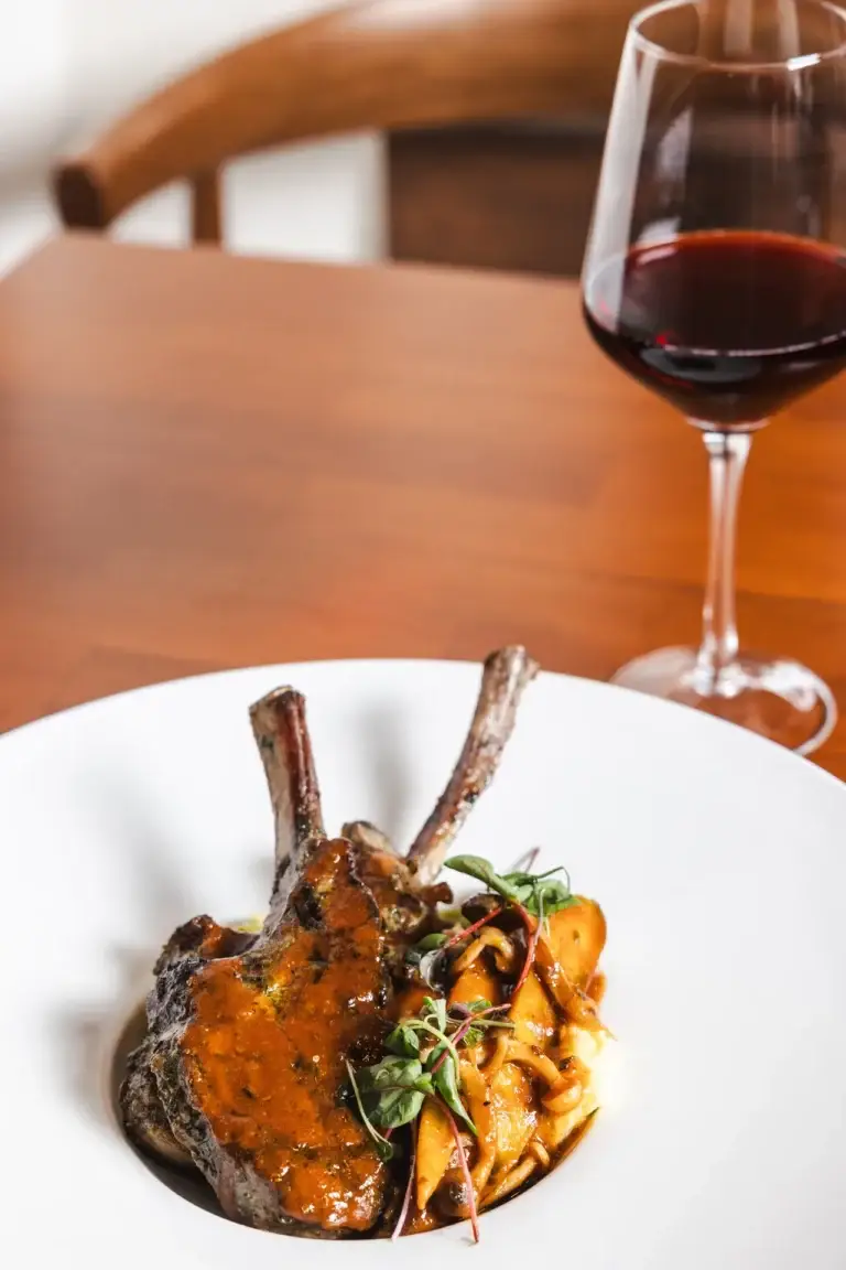 Wine & Bubbles 3 A glass of red wine behind Lamb Chops with sauce, carrots and mushrooms at Capriciano Osteria
