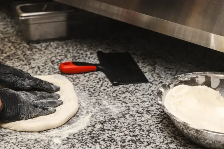 Gloved hands pressing out pizza dough on a floured granite counter at Capriciano Osteria