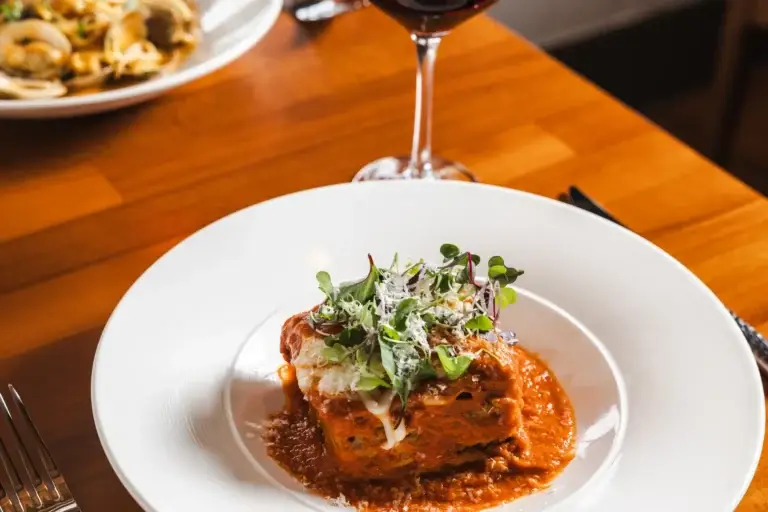 Layered lasagna in tomato sauce with microgreens and grated cheese, red wine behind, at Capriciano Osteria