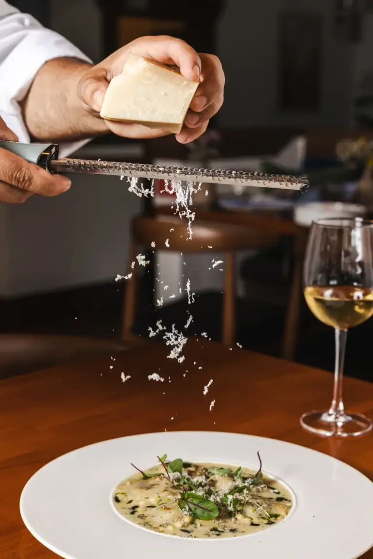 Parmigiano grated over a bowl of creamy ravioli, a glass of white wine nearby, at Capriciano Osteria