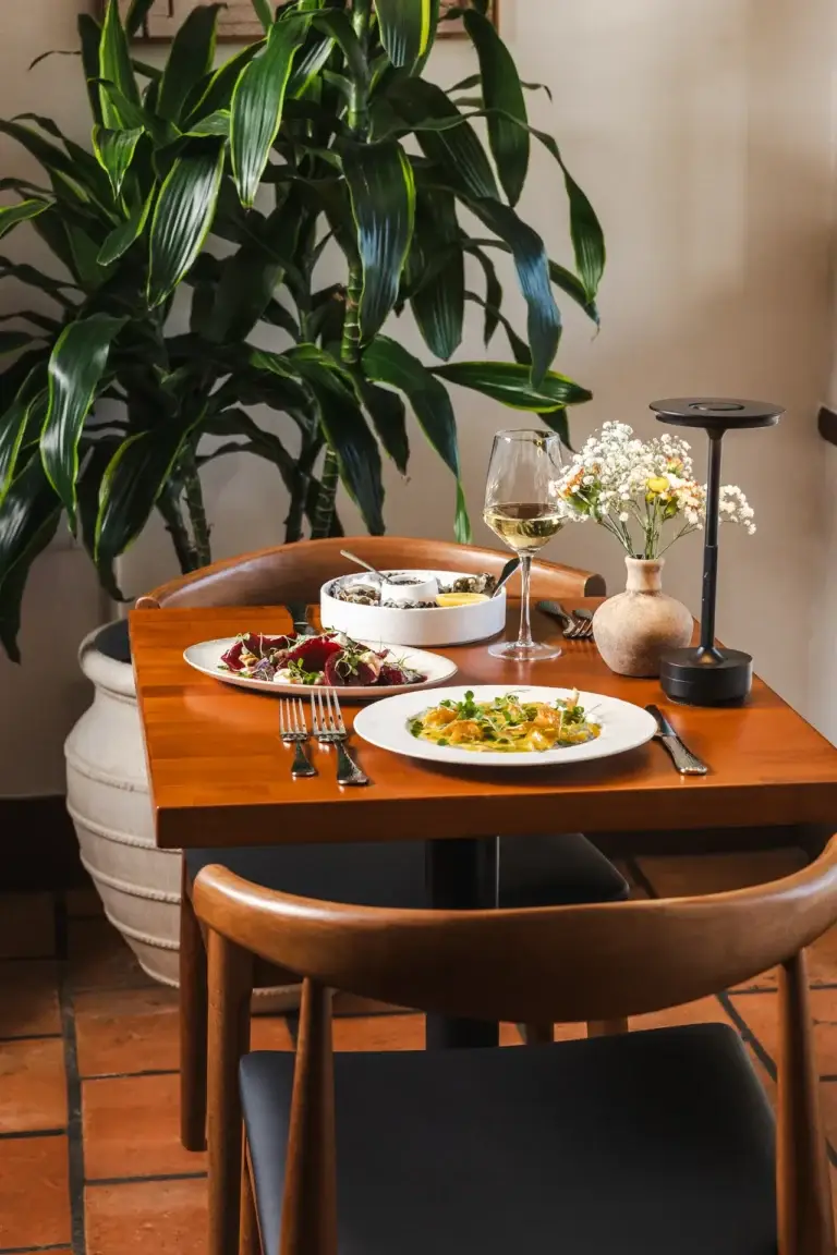 Make a Reservation 3 Table for two beside a large potted plant, set with plates, wine and flowers, at Capriciano Osteria